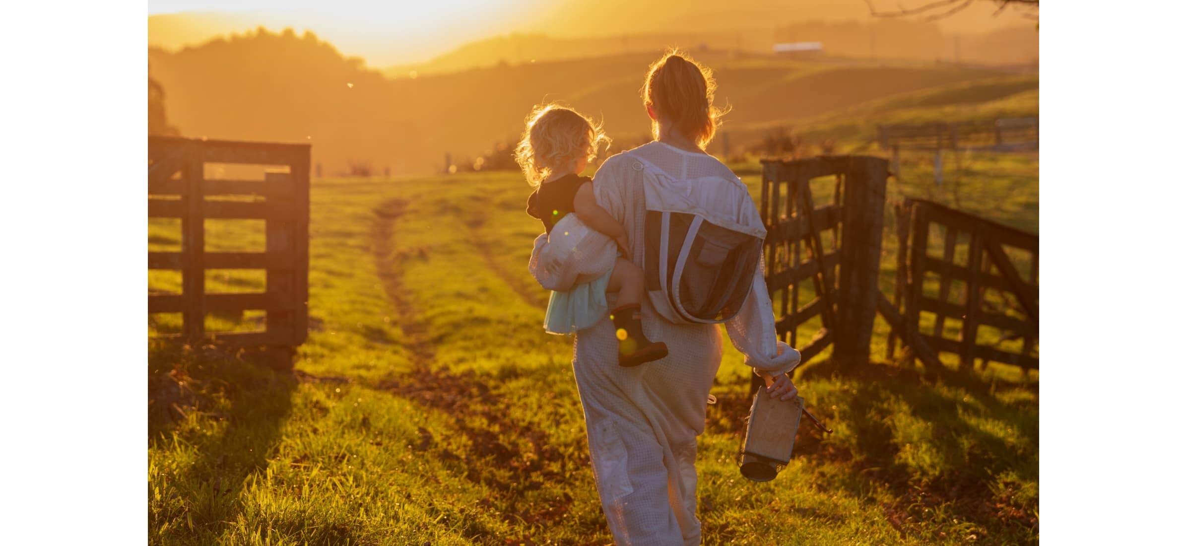 Hannah O’Brien walking through a farm gate at sunset, wearing a bee protection suit and carrying a toddler on her hip.